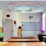 کلاس درس مدرسه‌ای با میزهای دانش‌آموزی، تابلوی سفید، کابینت‌های ذخیره‌سازی رنگی، آثار هنری دانش‌آموزان روی دیوار، و نورپردازی مناسب