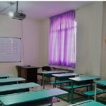 کلاس درس با میزهای دانش‌آموزی سبزرنگ، لوحه‌ی سفید، و ستائر بنفش روشن در دیواری که به نافذه می‌نگرد