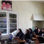 دانش‌آموزان در کلاس درس در حال شنیدن سخنرانی، فرد سخنران در سمت چپ با لپ‌تاپ، پوستر روی دیوار، قفسه‌های شیشه‌ای در پس‌زمینه