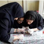 دو دانش‌آموز دختر با پوشش حجاب به رنگ مشکی در حال انجام یک آزمایش علمی در میز آزمایشگاه، با دستکش‌های پلاستیکی و مواد آزمایشی