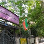 تابلوی ورودی مدرسه با نام‌نویسی پارسی، پرچم ملی ایران، و درب ورودی فلزی تزیینی
