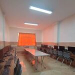 نمای داخلی سایت کامپیوتر مدرسه با ردیف‌های کامپیوتر رومیزی، صندلی‌های مشکی، میزهای کاری، و نور سقفی