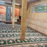 نمازخانه با فرش‌های سنتی سبز و صورتی، آیات قرآنی بر روی دیوارها، پنجره‌های پرده‌ای قرمز و نورپردازی طبیعی