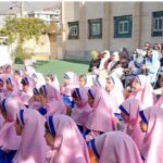 تجمع دانش‌آموزان با لباس یکدست صورتی‌رنگ در حیاط مدرسه به‌همراه حضور والدین و مسئولین برای شرکت در یک مراسم