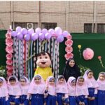 دانش‌آموزان دختری در لباس مدرسه صورتی و آبی در برابر بک‌دراپ تزئینی با گل‌های صورتی، بادکن‌های بنفش و عروسک کارتونی در حیاط مدرسه