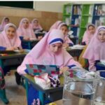 دانش‌آموزان دختری در حال انجام آزمایش علمی در کلاس درس، مشغول مشاهده و ثبت نتایج آزمایش با لیوان‌های آب