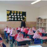 کلاس درس مدرسه ابتدایی با دانش‌آموزان دختر پوشنده حجاب صورتی نشسته در میز تحصیلی قرمز، معلم خانمی ایستاده، تابلو سیاه تزئین‌شده با نقاشی‌های خورشید و ابر، قفسه‌های خشبی در پس‌زمینه