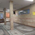 نمازخانه مدرسه با فرش‌های قرآنی، ستون‌های سفید، تزئینات اسلامی، و نورپردازی داخلی
