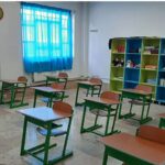 کلاس درس مجهز به میزهای ترسیمی سبز رنگ، صندلی‌های چوبی، قفسه‌های ذخیره‌سازی رنگی، پنجره‌های آبی و تابلوی دیواری هنری