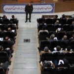 تصویری از یک مراسم اجتماعی داخل سالن با شرکت‌کنندگان نشسته روی صندلی‌های سیاه و یک سخنران ایستاده در میانه‌ی سالن زیر بنر فارسی