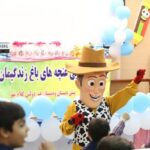 دانش‌آموزان در حال تماشای یک برنامه تئاتری با کارکتر بزرگ و تزئینات رنگین و بنرهای فارسی در سالن مدرسه