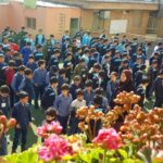 گروهی از دانش‌آموزان در حیاط مدرسه در کنار گل‌های رنگی و ساختمان‌های مدرسه