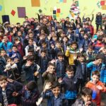 عکس گروهی از دانش‌آموزان ایستاده در مقابل دیوار تزئین‌شده با کاغذهای رنگی و نقاشی در جریان یک رویداد مدرسه‌ای