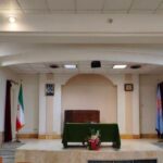 نمای داخلی نمازخانه مدرسه با منبر چوبی، پرچم ایران، و میز سبز رنگ برای نماز