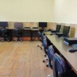 اتاق کامپیوتر مدرسه با میزهای تیره، صندلی‌های مشکی و رایانه‌های رومیزی در کنار دیوار