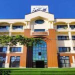 نمای خارجی ساختمان مدرسه با معماری معاصر، سردرِ ورودی، تابلوی شناخت‌نامه، درب‌های متعدد، و درختان سبز