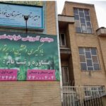 نمای بیرونی ساختمان خاکی‌رنگ مدرسه با تابلوی آموزشی، پنجره‌های متقارن، و درب ورودی فولاد‌ی