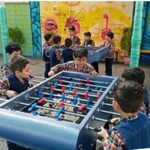 دانش‌آموزان در حال بازی با فوتبال دستی در حیاط مدرسه، جوانان با پیراهن کارو رنگی در حال تمرین و تفریح