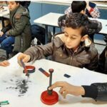 گروهی از دانش‌آموزان در حال انجام یک فعالیت عملی و آزمایش علمی با استفاده از تجهیزات آزمایشگاهی، شامل مغناطیس و ابزارهای دقیق