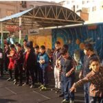 دانش‌آموزان کوچک در حیاط مدرسه زیر سقف پوشیده ایستاده‌اند و برچسب‌های رنگی روی دیوار پس‌زمینه قابل دید است