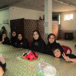 دسته‌ای از دانش‌آموزان دختر حول یک میز در یک برنامه گروهی پیش‌رو، برخی نشسته و برخی ایستاده، با تزیین قرمز رنگ بر روی میز و تخته‌های نمایشی در پس‌زمینه