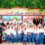 عکس گروهی دانش‌آموزان و معلمان ایستاده در مقابل دیوار رنگی با نام مدرسه خجستگان زهرا