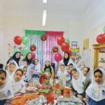 گروهی از دانش‌آموزان دختر در حال برگزاری مراسم جشن در کلاس درس با تزئینات بادکنک رنگی، پارچه‌های سبز و قرمز، و میز جشن
