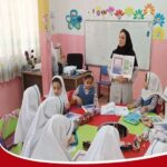 معلمی در حال نشان‌دادن کتاب و اسلاید آموزشی به دانش‌آموزانی که دور میز سبز و مواد درسی نشسته‌اند، در کلاس درسی با دیوار‌های صورتی و تزئینات رنگی