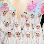 گروهی از دانش‌آموزان دختر با لباس و حجاب سفید ایستاده در حین حضور در یک جشن یا رویداد مدرسه‌ای با پس‌زمینه تزئین شده به گل‌های کاغذی و تزئینات رنگی
