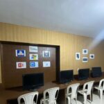 اتاق سایت کامپیوتر مدرسه با میز‌های کاری، مانیتورهای کامپیوتری، صندلی‌های سفید و سیاه، و دیوار دکوراتیو با لوگوهای نرم‌افزاری