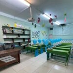 نمایی داخلی از کلاس درسی با میز‌های سبز، صندلی‌های آبی، قفسه‌های کتاب، و تزیینات آموزشی.