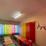 کلاس درس کودکان با میزهای رنگی، صندلیهای رنگین، دیوارهای صورتی و زرد، تزیینات کودکانه و پرده‌های رنگی