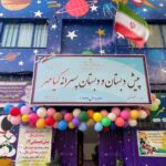 نمای سردرِ مدرسه با تابلو، بادکنک‌های رنگی و پرچم ایران