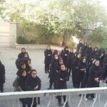 گروهی از دانش‌آموزان با لباس سیاه در حیاط مدرسه؛ فضای باز با دیوار، دروازه و درختان سبز