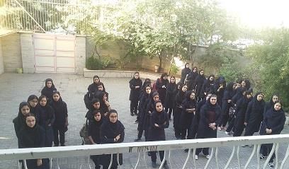 گروهی از دانش‌آموزان با لباس سیاه در حیاط مدرسه؛ فضای باز با دیوار، دروازه و درختان سبز