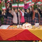گروهی از دانش‌آموزان دختر در حیاط مدرسه که در مراسم جشن میلاد جمهوری اسلامی ایران، پرچم‌های ملی را در دست گرفته‌اند و بر سر میز جشن با کیک و دکوراسیون سه‌رنگی ایستاده‌اند.