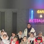 دسته‌ای از دانش‌آموزان در کنار یک نیون ساین برد طراحی شده با متن کافه رستوران در حال پوز برای عکس یادگاری