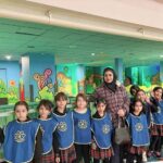 گروهی از دانش‌آموزان و معلمی در راهروی مدرسه، ایستاده در کنار دیوار تزئین‌شده با نقاشی‌های رنگین گلها و کاکتوس، برای عکس یادگاری یادی.
