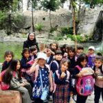گروهی از دانش‌آموزان با معلم در حال بازدید از باغ وحش یا منطقه طبیعی حفاظت‌شده، در کنار آبشار و محیط سنگی