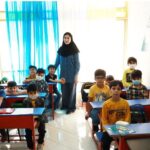 معلم و دانش‌آموزان در فضای کلاس درس روشن با میزهای قرمز و آبی و پنجره‌های پرده‌دار