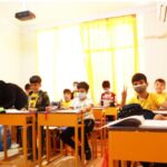 کلاسی پر از دانش‌آموزان در حال درس و یادگیری، معلم در کنار میزهای نارنجی و دیکوریشن آموزشی زرد