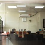کلاس درسی با نورپردازی داخلی، دانش‌آموزان در حال فعالیت پشت میزها، تابلوهای دیواری، گل‌های درون‌فضایی و قفسه‌های ذخیره‌سازی