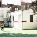 حیاط مدرسه با سطح سبز رنگ، دیوارهای سفید و خطوط سرخ، ساختمان‌های آجری در پس‌زمینه و دانش‌آموزان در حال بازی