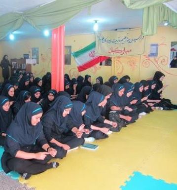 گروهی از دانش‌آموزان دختر نشسته در فضای داخلی سالن مدرسه در حال شرکت در یک مراسم یا فعالیت گروهی