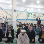 گروهی از دانش‌آموزان و معلمان در حال شرکت در نمازِ جماعت در سالن‌ داخلی مدرسه با تزئینات دیواری و تابلوهای دسته‌جمعی