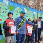 دانش‌آموزان و مسئولین مدرسه در حال شرکت در مراسم تقدیری و جشن تشریفات با بنر تزیین شده در محوطه بیرونی
