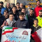 گروهی از دانش‌آموزان و بزرگسالان در یک مراسم فرهنگی درباره تاریخ انقلاب اسلامی، با نوار اطلاعات و پرچم ایران