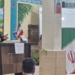 مردی در کت‌وشلوار در حال سخنرانی رسمی در مدرسه، میکروفن در پشت سخنگو، پرچم‌های ایران و استانی در پس‌زمینه