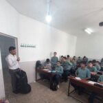 نمای داخلی کلاس درس با معلم و دانش‌آموزان نشسته بر روی نیمکت‌ها در حال تدریس