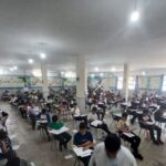 دانش‌آموزان در حال انجام امتحان در کلاس درسی با میزهای فردی و صندلی‌های سیاه، با تابلوهای یادآوری و تزئینات دیواری